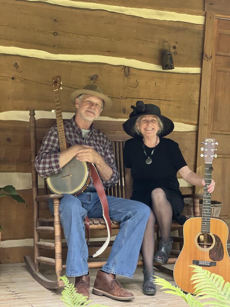 Dale Farmer and Ma Crow of Farmer and the Crow sitting on a front porch rocking chair, holding a guitar and banjo.